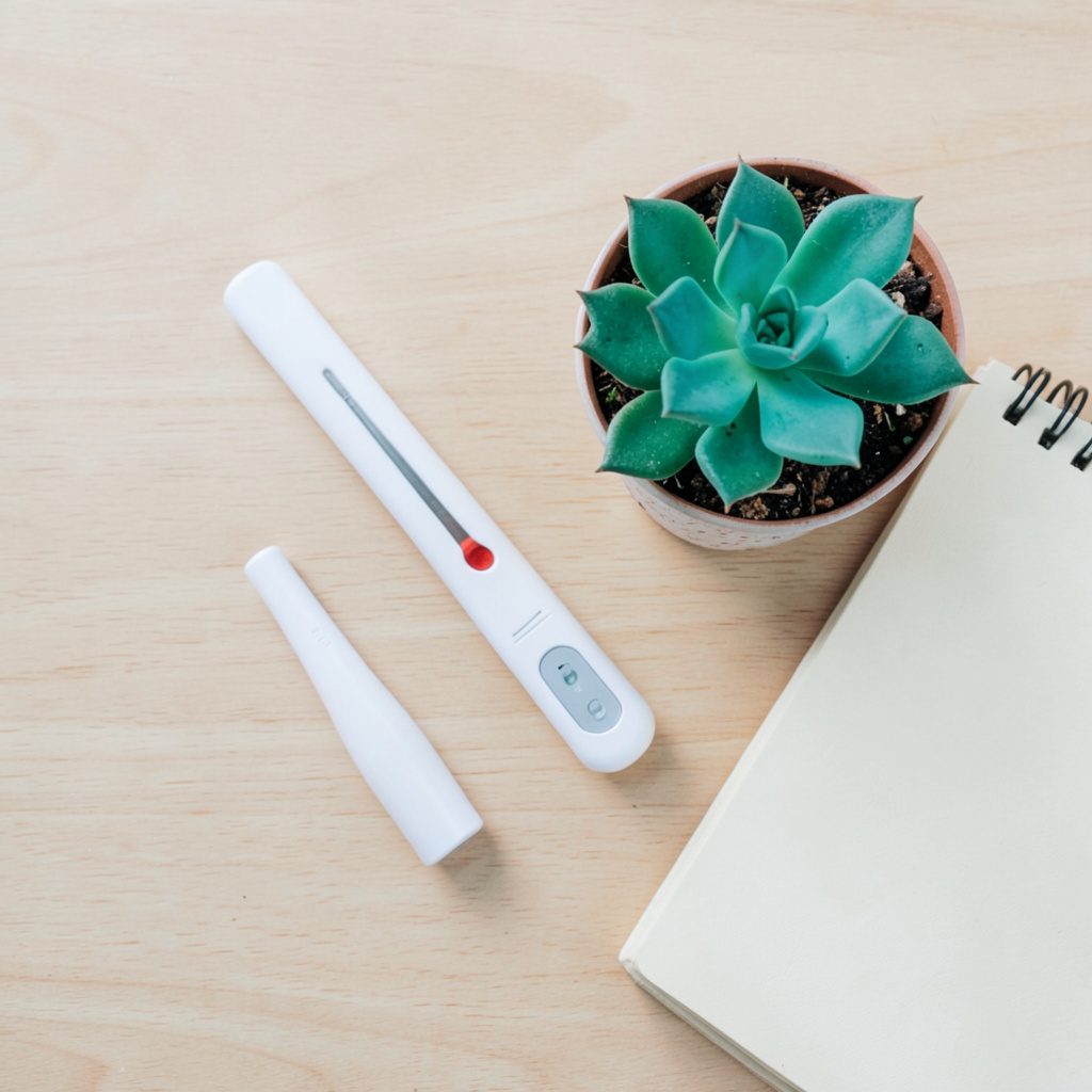 Overhead flat-lay of a compact white thermometer and humidity sensor on a light wooden surface, beside a small potted succulent and a notebook