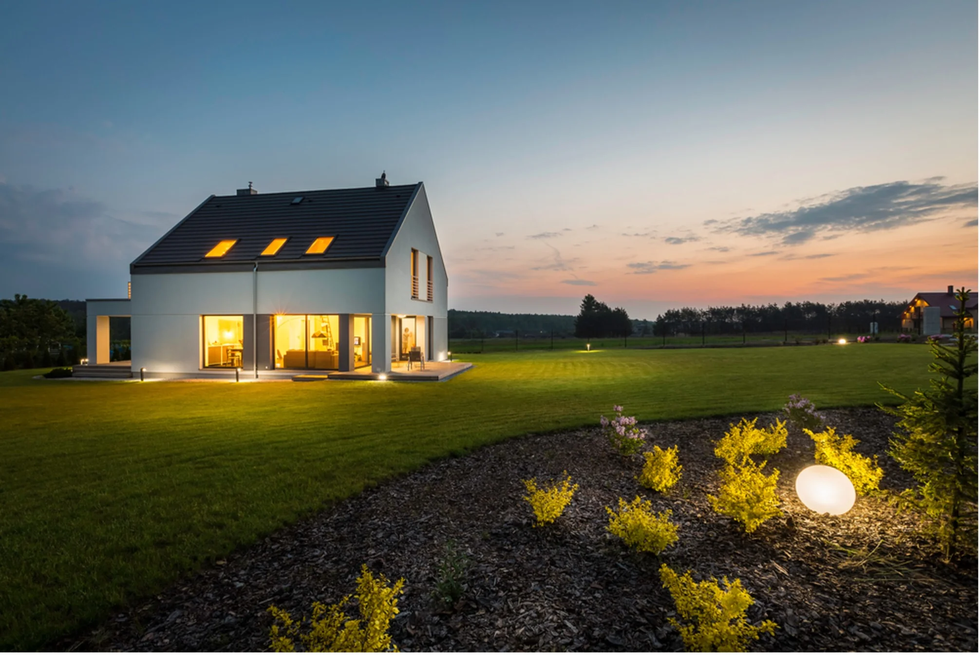 House exterior at dusk with warmly lit windows, a neat lawn and glowing garden lights among yellow flowers