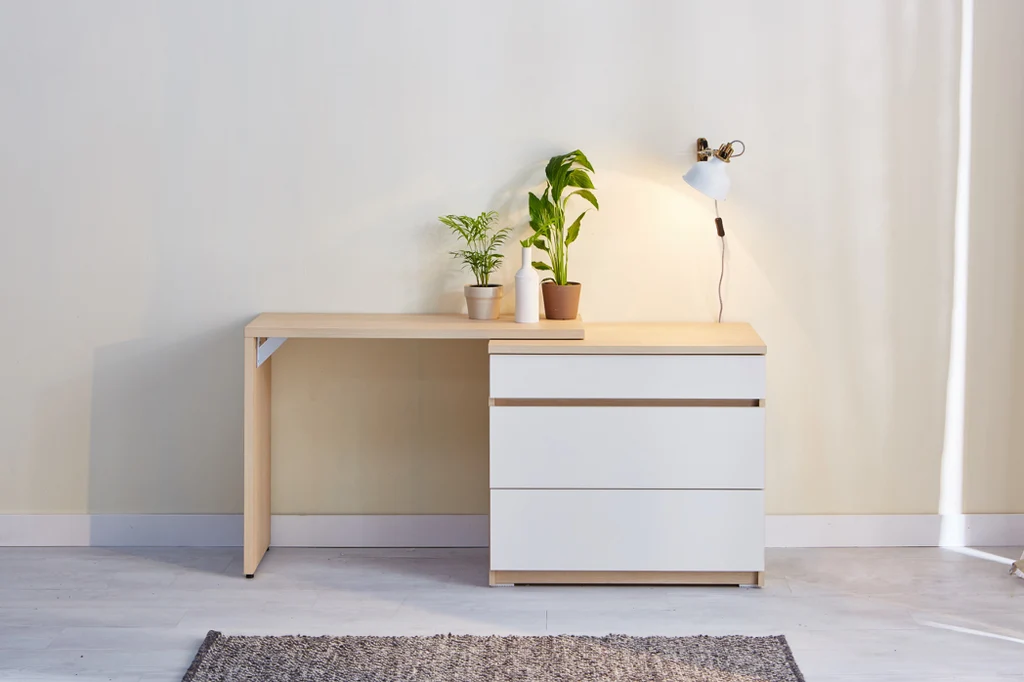 Overhead flat-lay photograph of a simple wooden desk with white drawers, two small potted plants, a compact wall lamp and a folded rug on light flooring