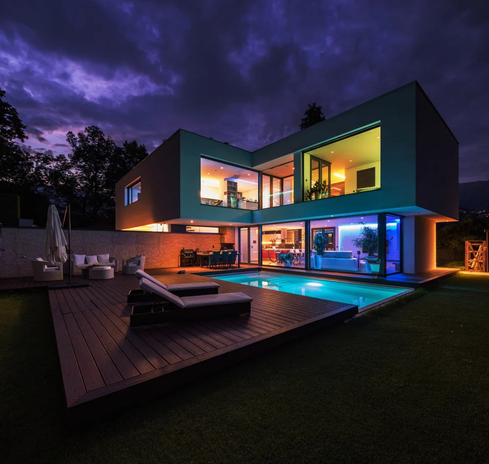 Modern two-storey home at night with warm and colourful interior lighting glowing through large windows, a still pool reflecting the lights and a wooden deck with lounge chairs