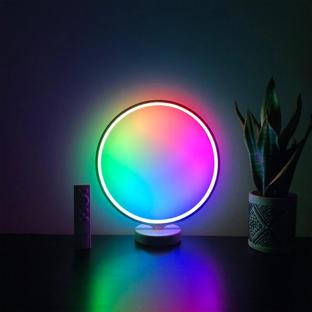 Round ring light glowing in rainbow colours on a dark tabletop beside a small remote and a potted plant, moody low-key scene